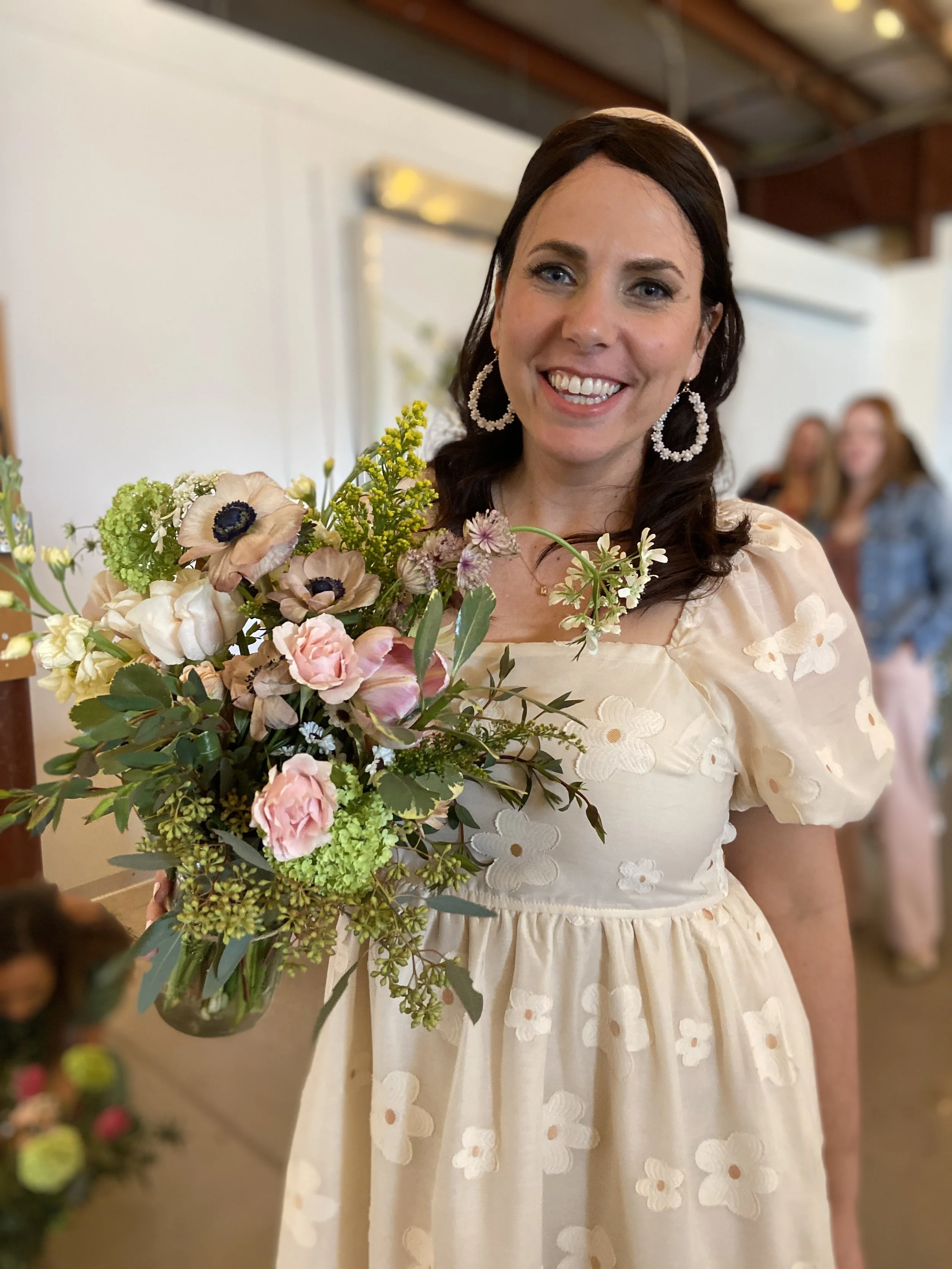 A woman with dark brown hair smiling and holding a large bouquet of pink, cream, and purple flowers. She wears a cream-colored dress with floral embroidery and hoop earrings. Blurred people are visible in the background.