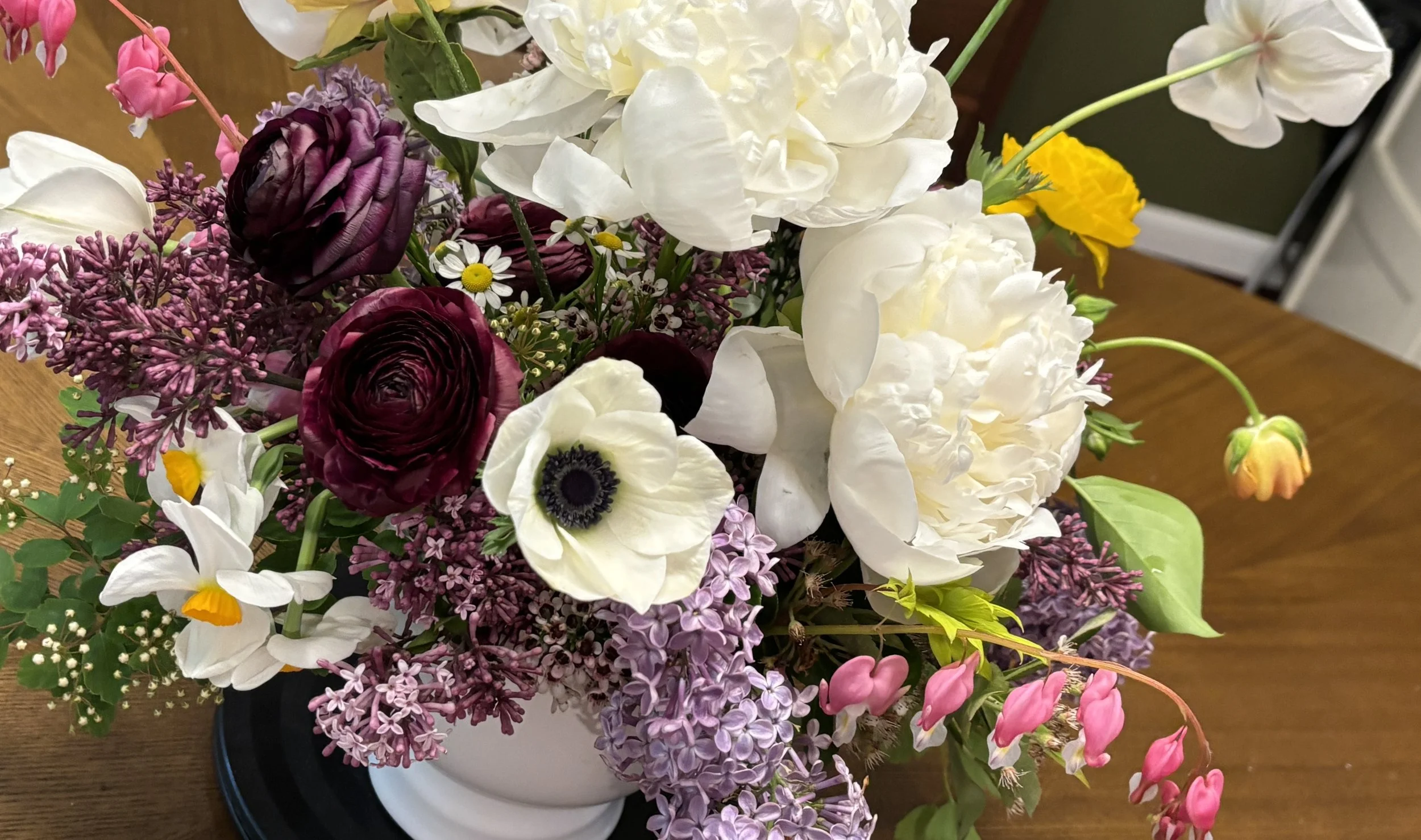 A colorful flower bouquet including white anemones, deep purple ranunculus, pink and white lilies, yellow and white daisies, and purple lilacs, arranged in a white vase on a wooden table.