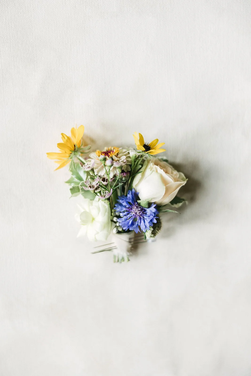 Small bouquet of mixed flowers including white roses, yellow, purple, and blue flowers, arranged in a small glass container on a light-colored background.