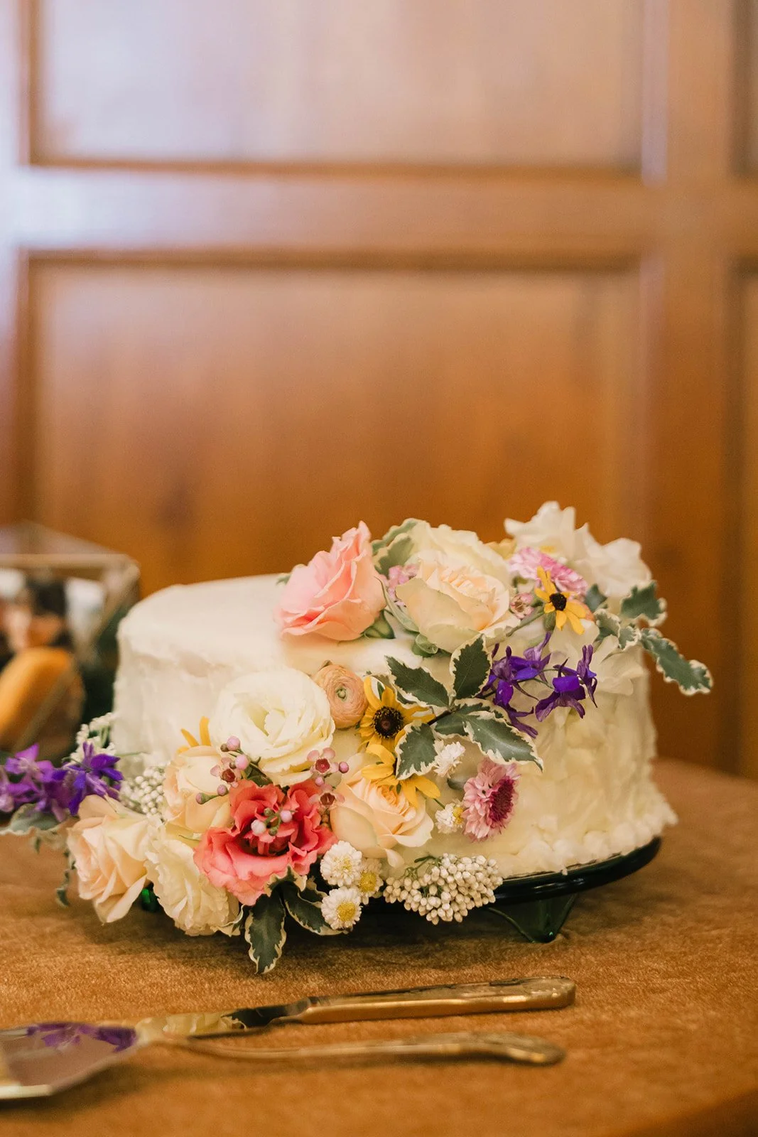A small white frosted cake decorated with pink, white, purple, and yellow flowers, sitting on a wooden table with a candlestick and cake knife nearby.