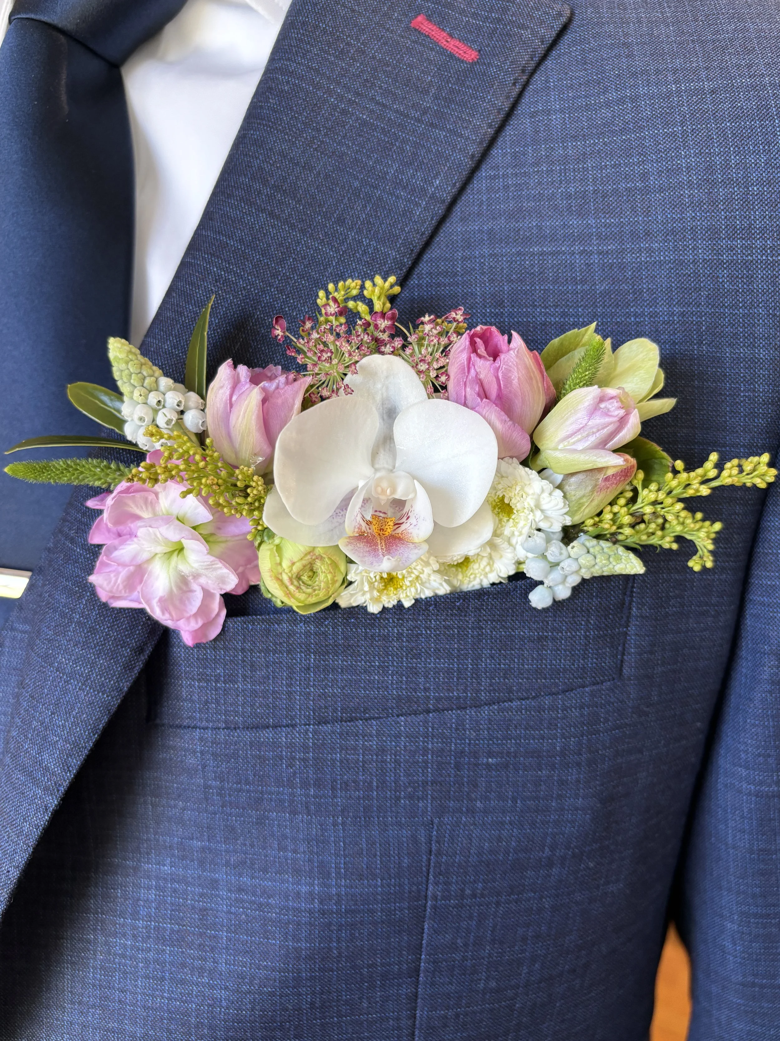 A close-up of a men's dark blue textured suit jacket with a pink stitch detail near the lapel, worn with a white shirt and a dark blue tie. A floral boutonniere with pink and white flowers and green leaves is pinned to the lapel.