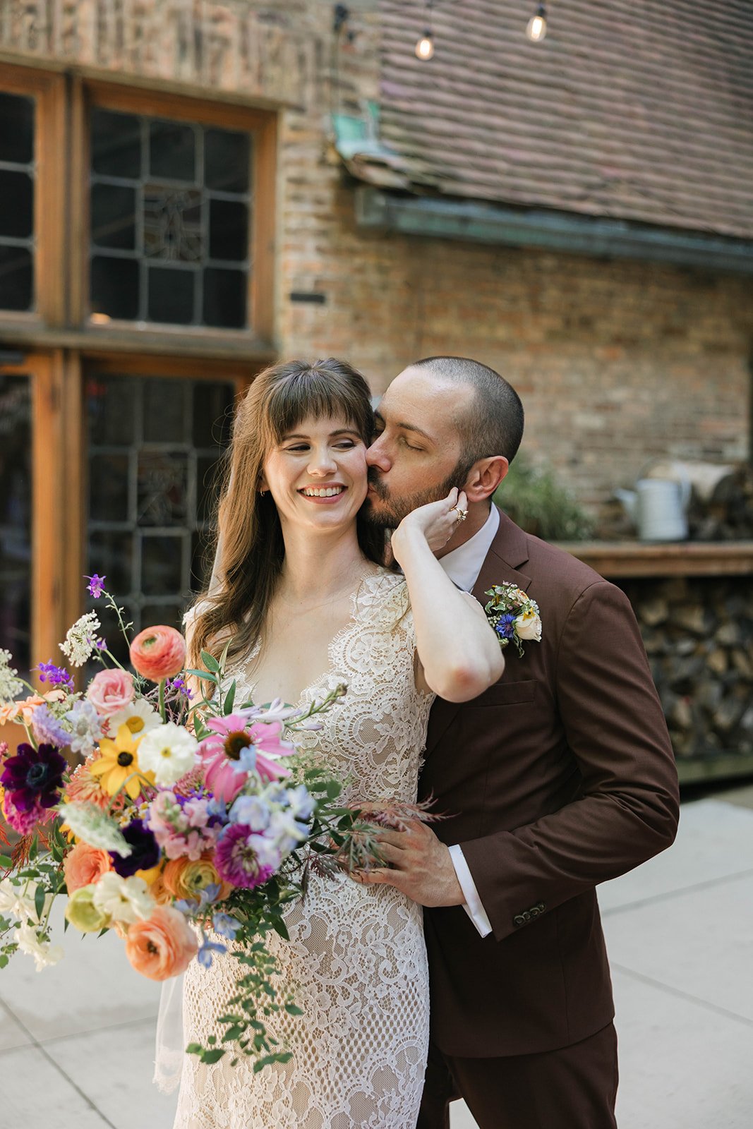 A bride and groom embracing outdoors, with the groom kissing the bride's cheek. The bride is holding a colorful bouquet of flowers, and the background shows a brick building and wood logs.