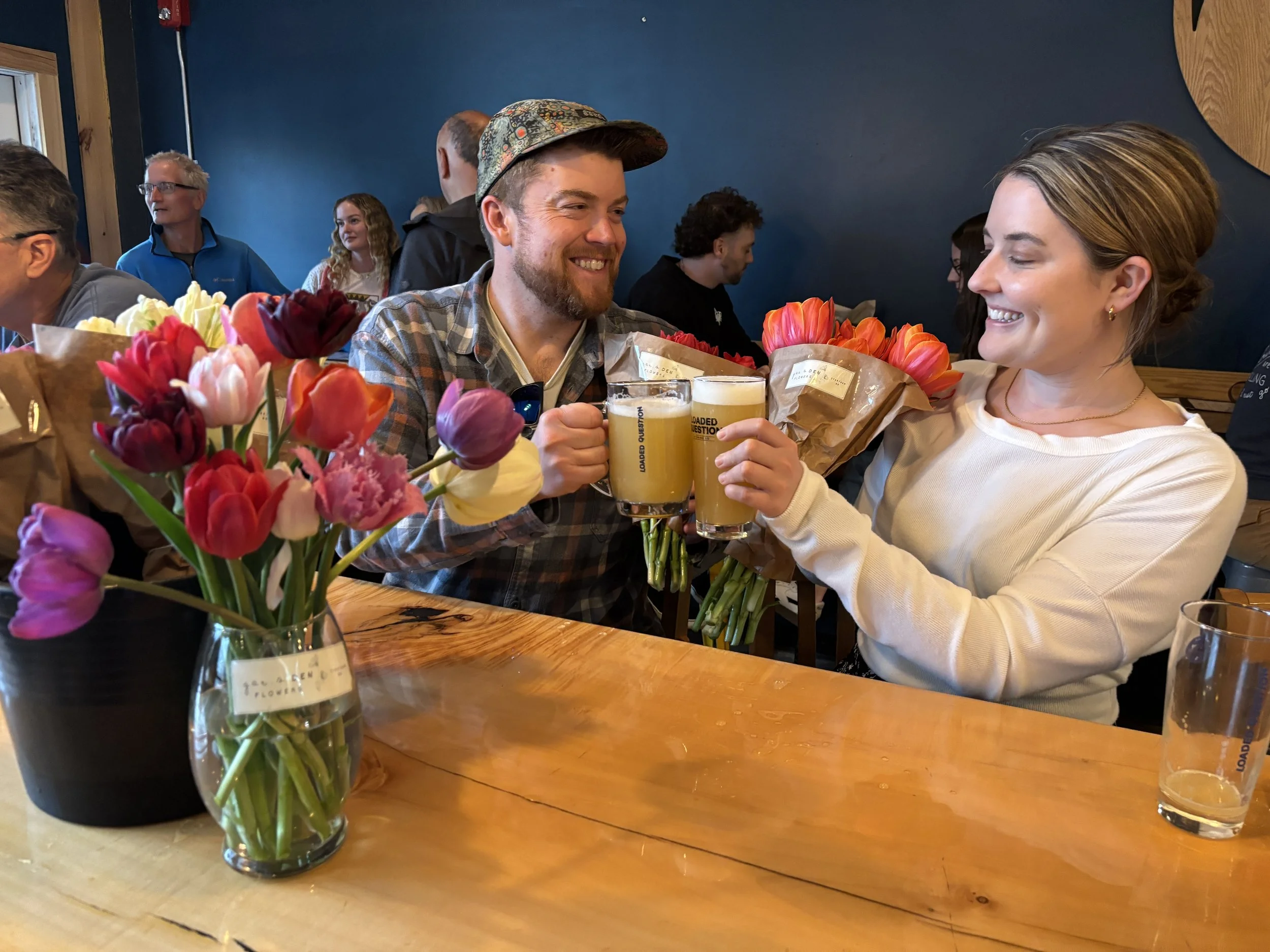 A group of people sitting at a wooden bar at the brewery Loaded Question, smiling and holding glasses of beer. There are bouquets of colorful tulips on the table, and a blue wall in the background.