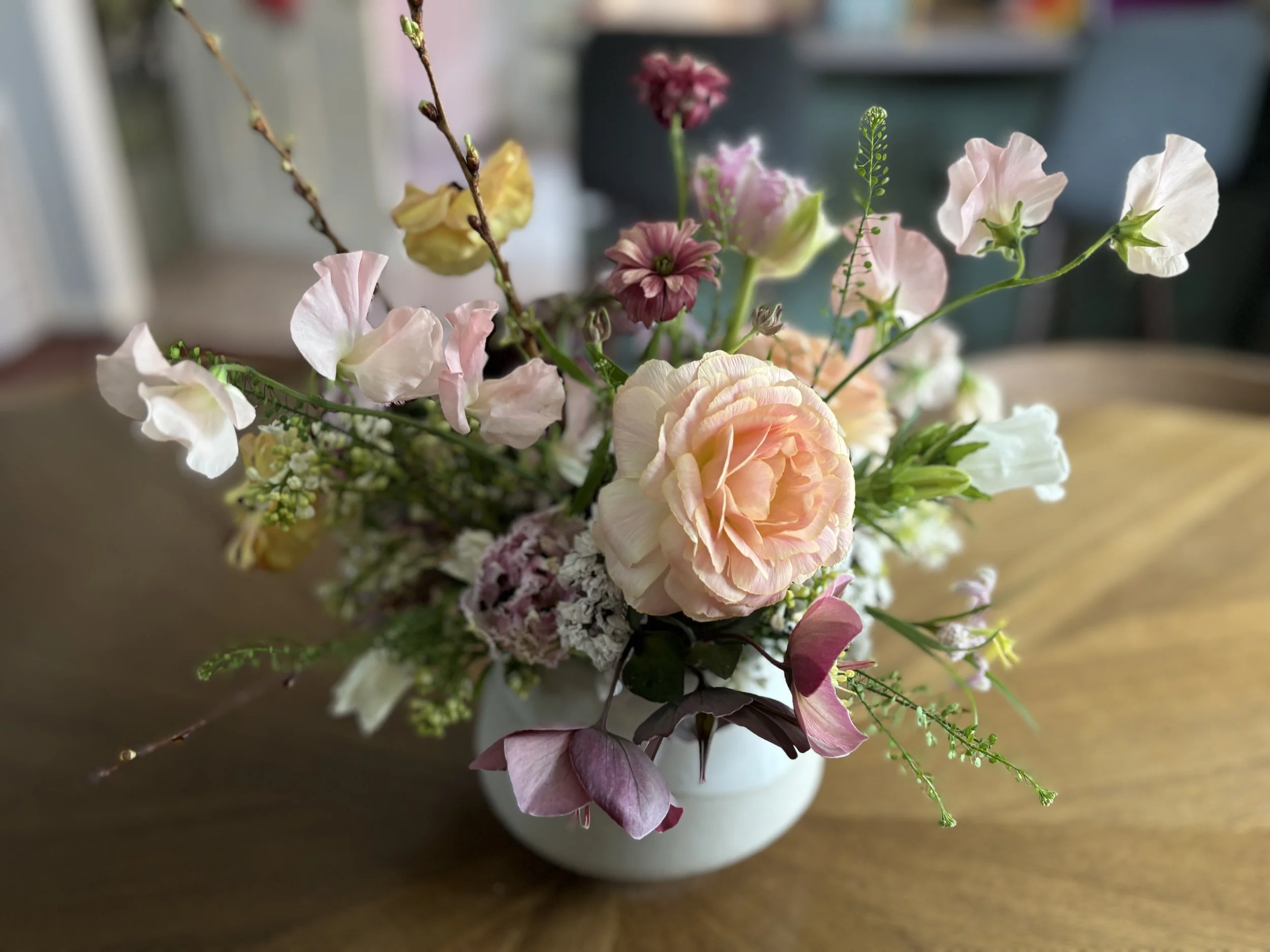 A bouquet of pastel-colored flowers including pink roses, white and blush blossoms, green foliage, and some sprigs of small white filler flowers, arranged in a white vase on a wooden surface.
