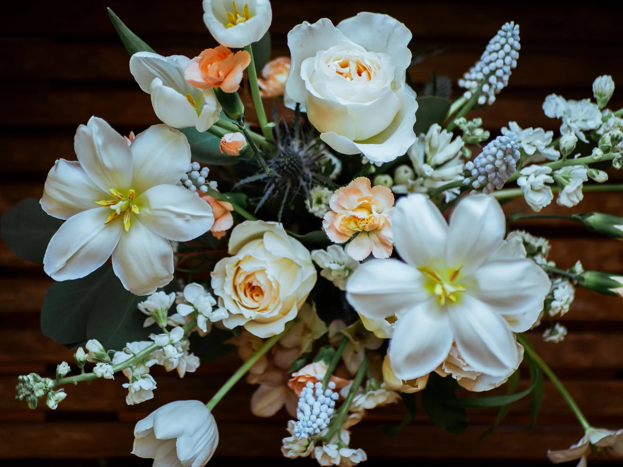 A bouquet of white and peach flowers with green leaves on a dark wooden background.
