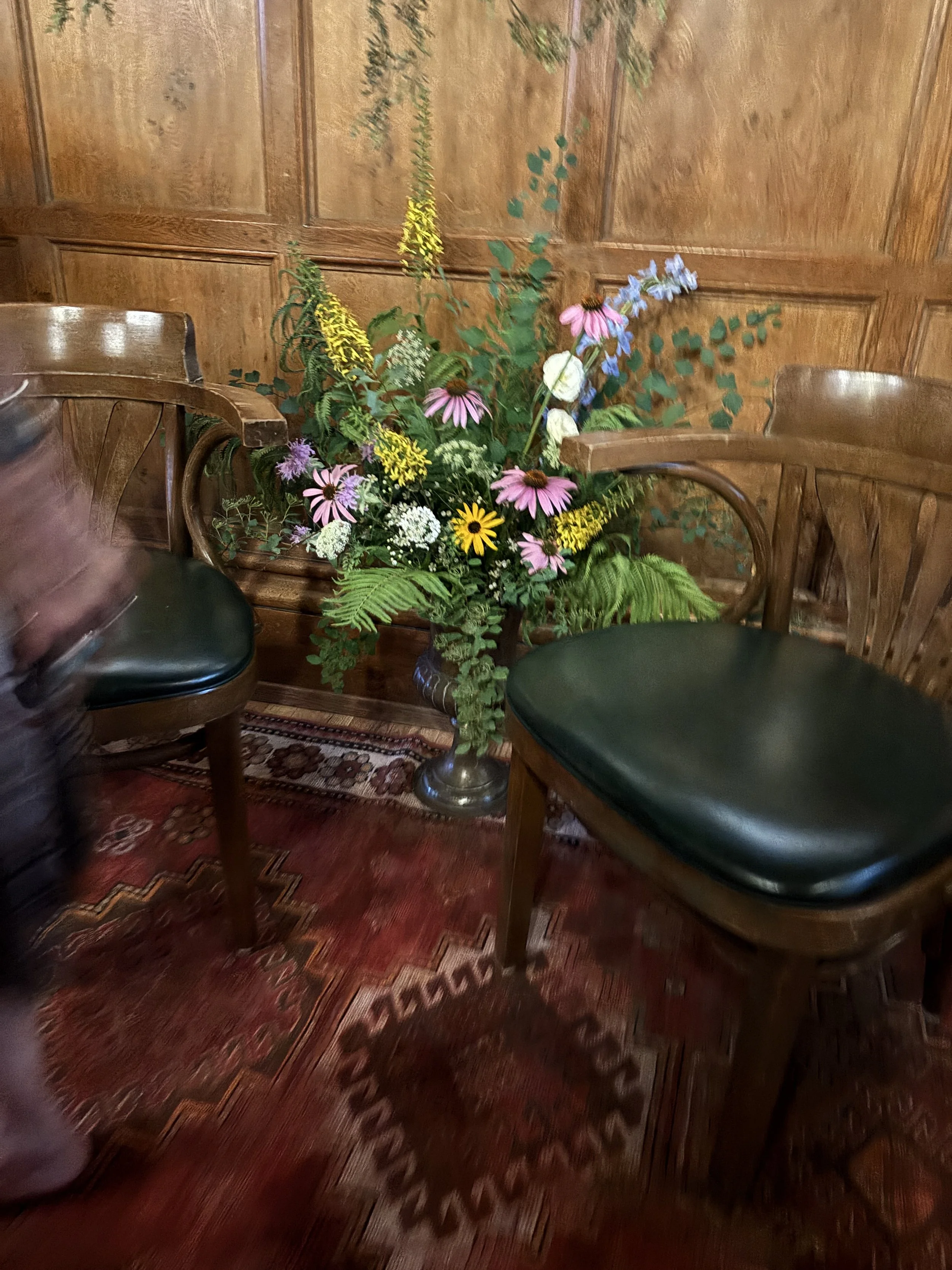 A wooden-paneled wall with a flower arrangement in a pot, surrounded by two vintage wooden chairs with green leather seats, on a red patterned rug.