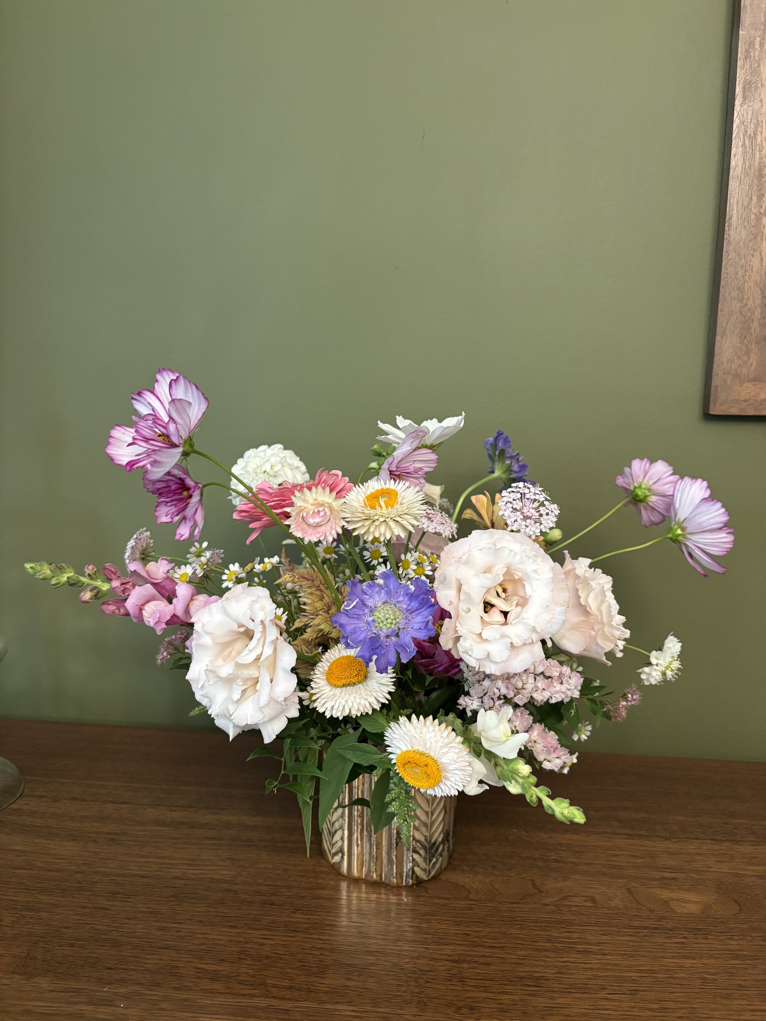 A colorful bouquet of mixed flowers in a rustic metal vase on a wooden table against a green wall.