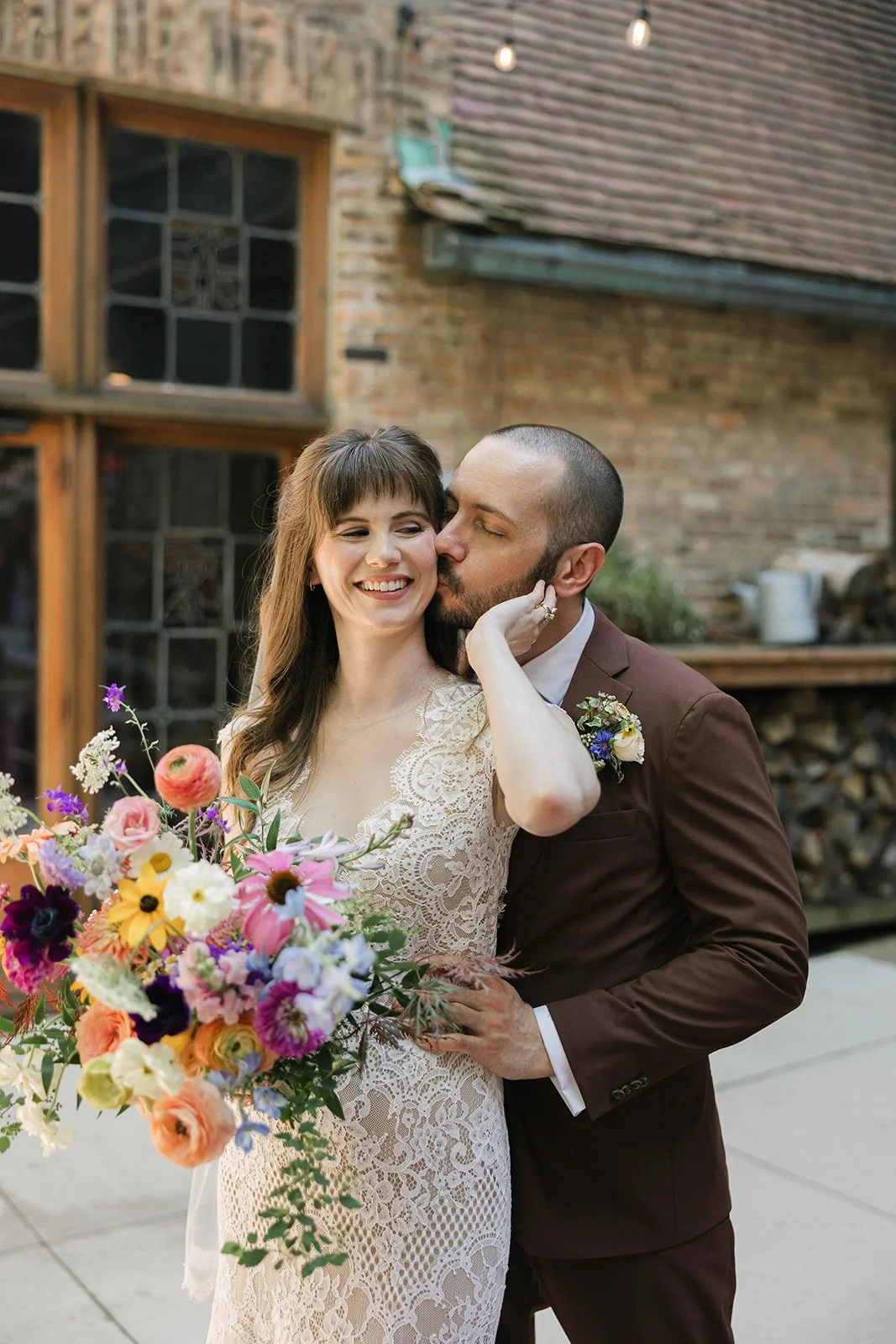 A newlywed couple shares a kiss outdoors, with the groom kissing the bride's cheek while she smiles and holds a colorful bouquet of flowers.