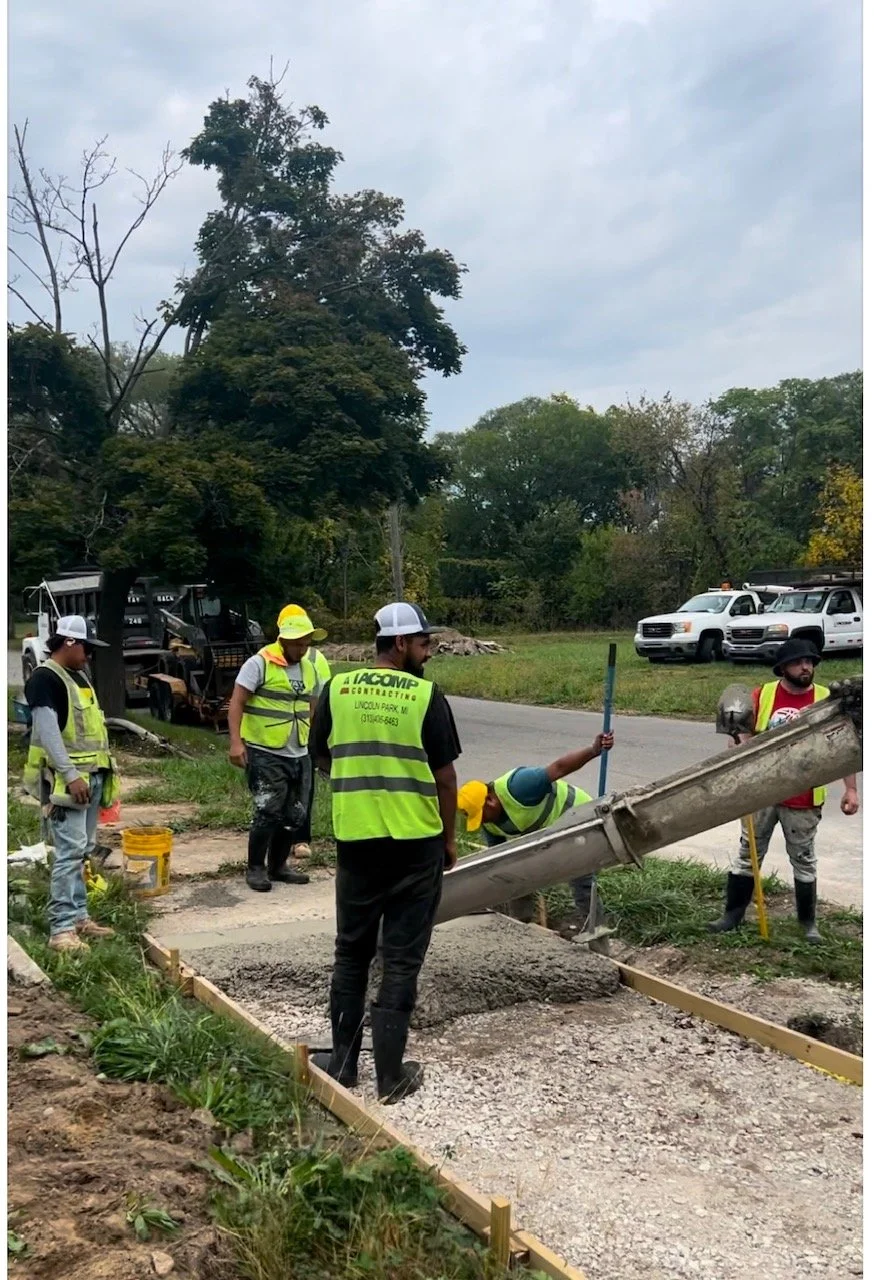 Construction workers are pouring concrete on a sidewalk, with some holding tools and wearing safety gear, surrounded by vehicles and trees.