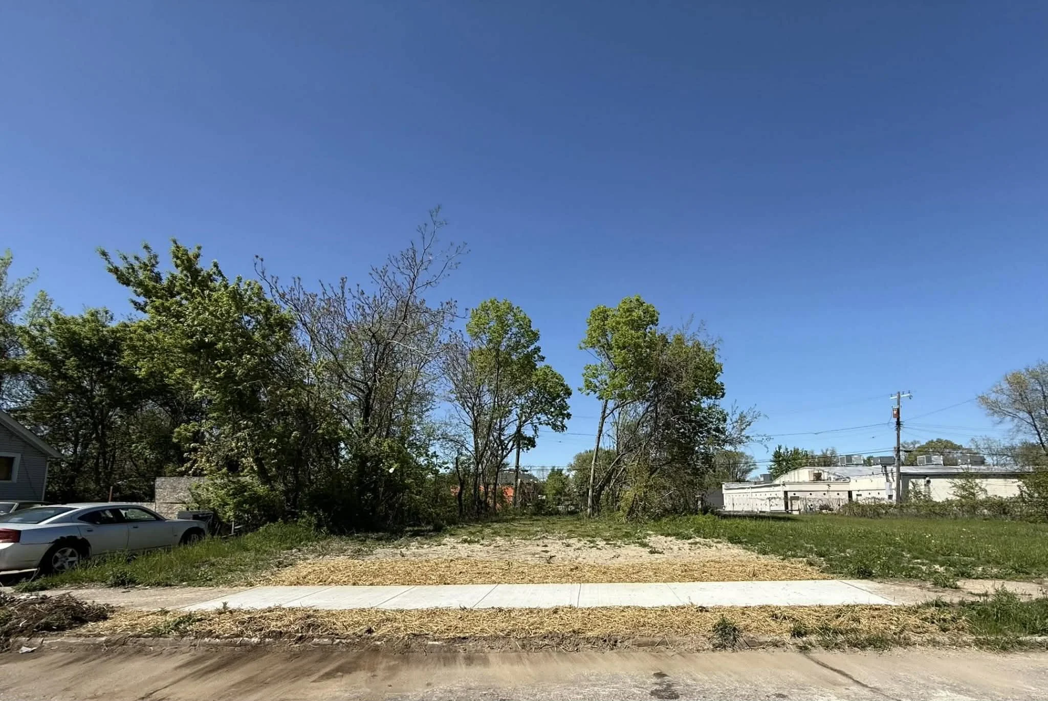 An empty lot with a sidewalk, some grass, and trees in the background under a clear blue sky.