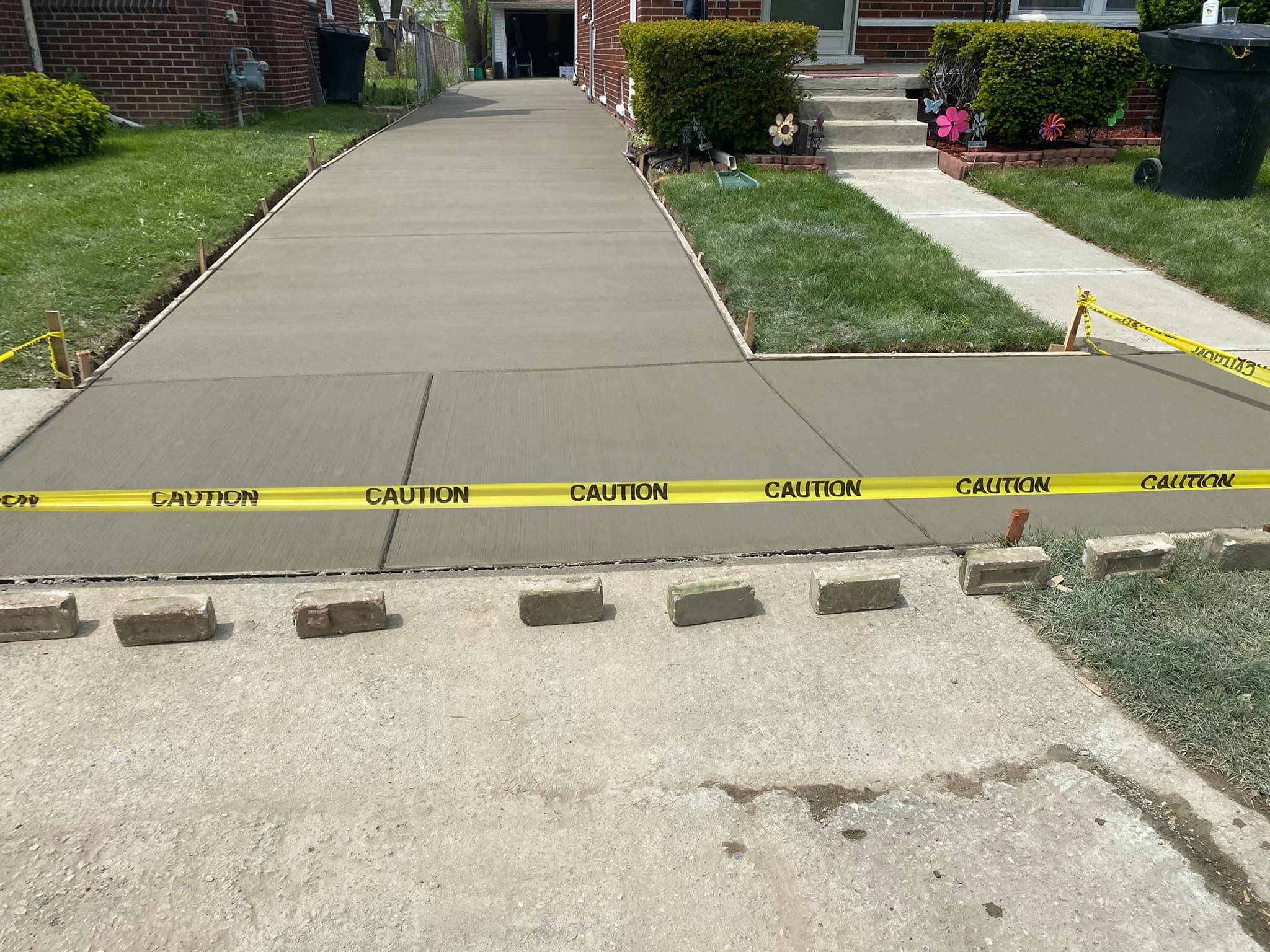 Freshly poured concrete sidewalk section with yellow caution tape marking the area, and wooden and concrete blocks placed around the edges for protection or to prevent access.