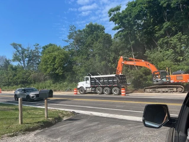 Construction work on a highway with an orange excavator, a white dump truck, and orange traffic cones. Traffic is stopped, and the view is from inside a vehicle.