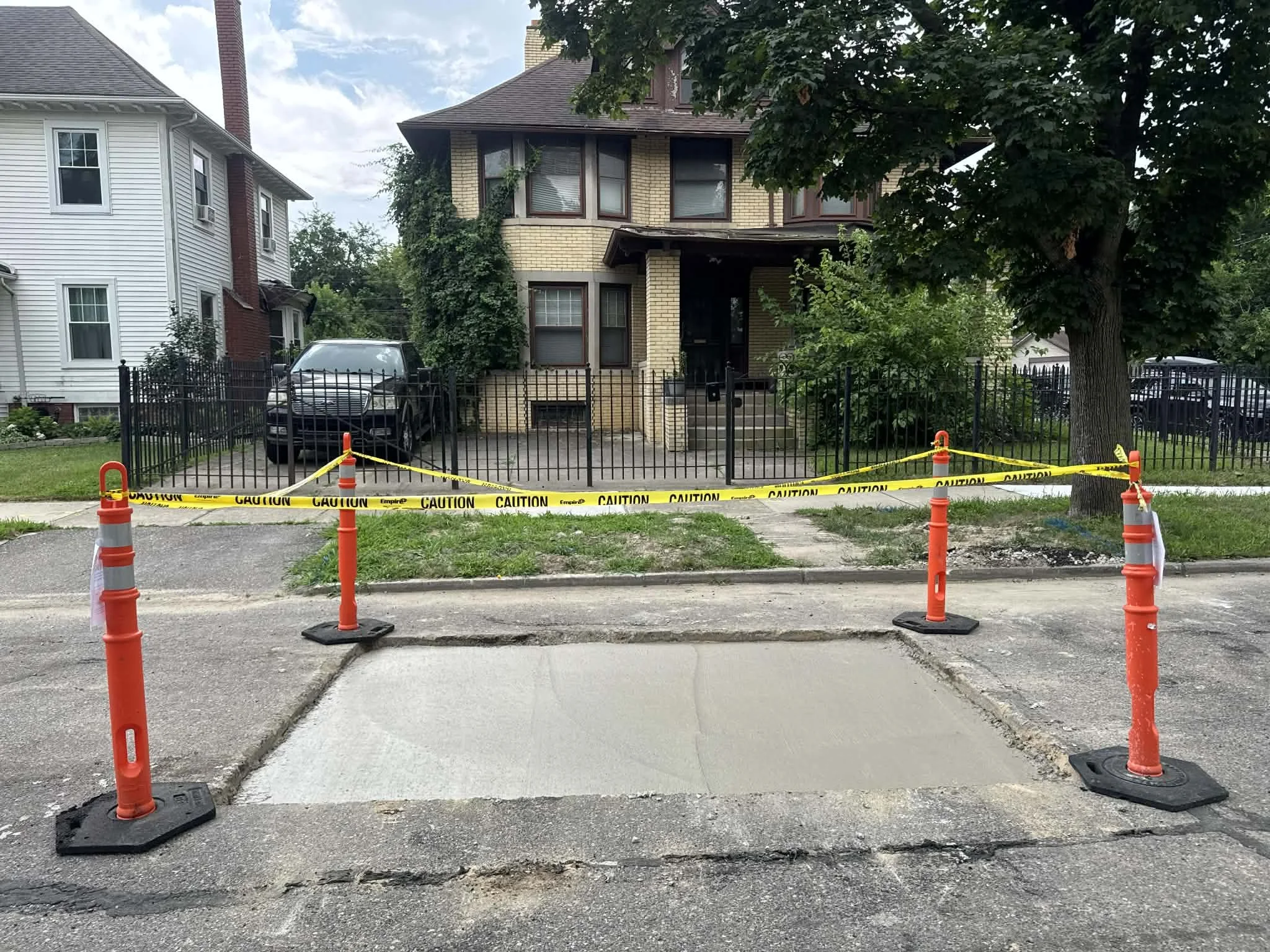 Street view of a sidewalk with a section under construction, surrounded by orange cones and yellow caution tape. In the background, there is a house with a brick facade, some greenery, and a parked car.
