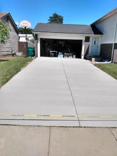 Clean concrete driveway leading to an open garage with outdoor furniture inside, adjacent to a house with a basketball hoop in the backyard, and a blue sky overhead.