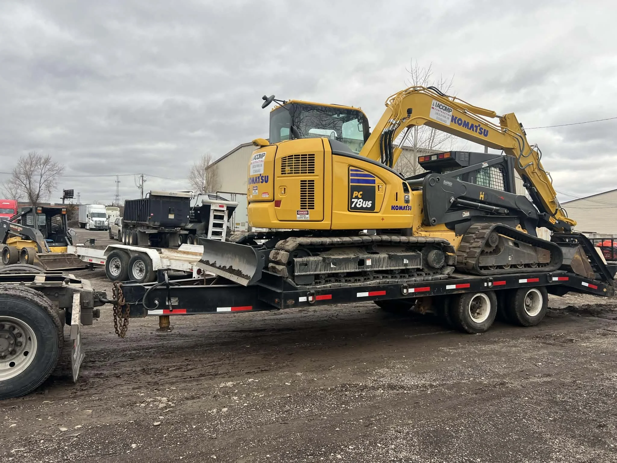 A yellow Komatsu PC78US excavator on a flatbed trailer in a construction yard, with other construction equipment and trucks in the background under cloudy skies.