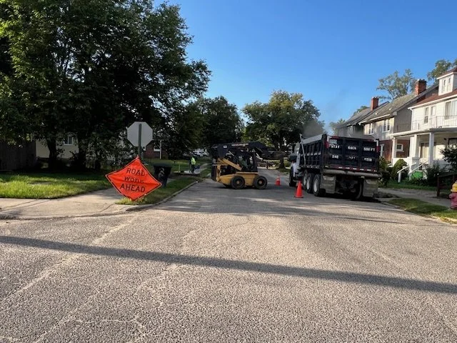 Construction vehicles and equipment blocking a residential street with an orange 'Road Work Ahead' sign and traffic cones.