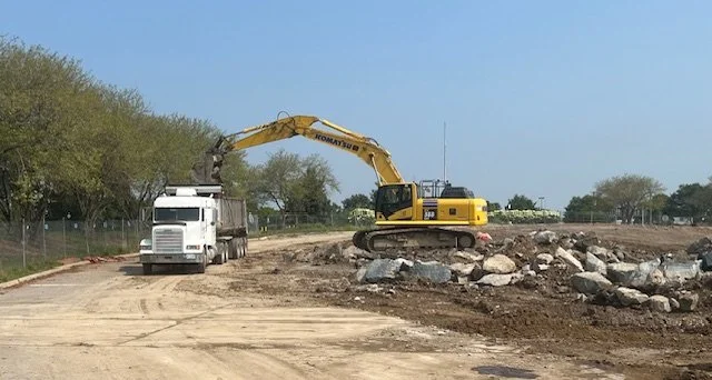 Construction site with an excavator loading dirt into a dump truck, with rocks and dirt on the ground and trees in the background.