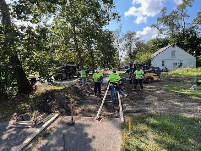 Workers in safety vests excavating and working on a construction site near a residential area, with trees, vehicles, and a white house in the background.
