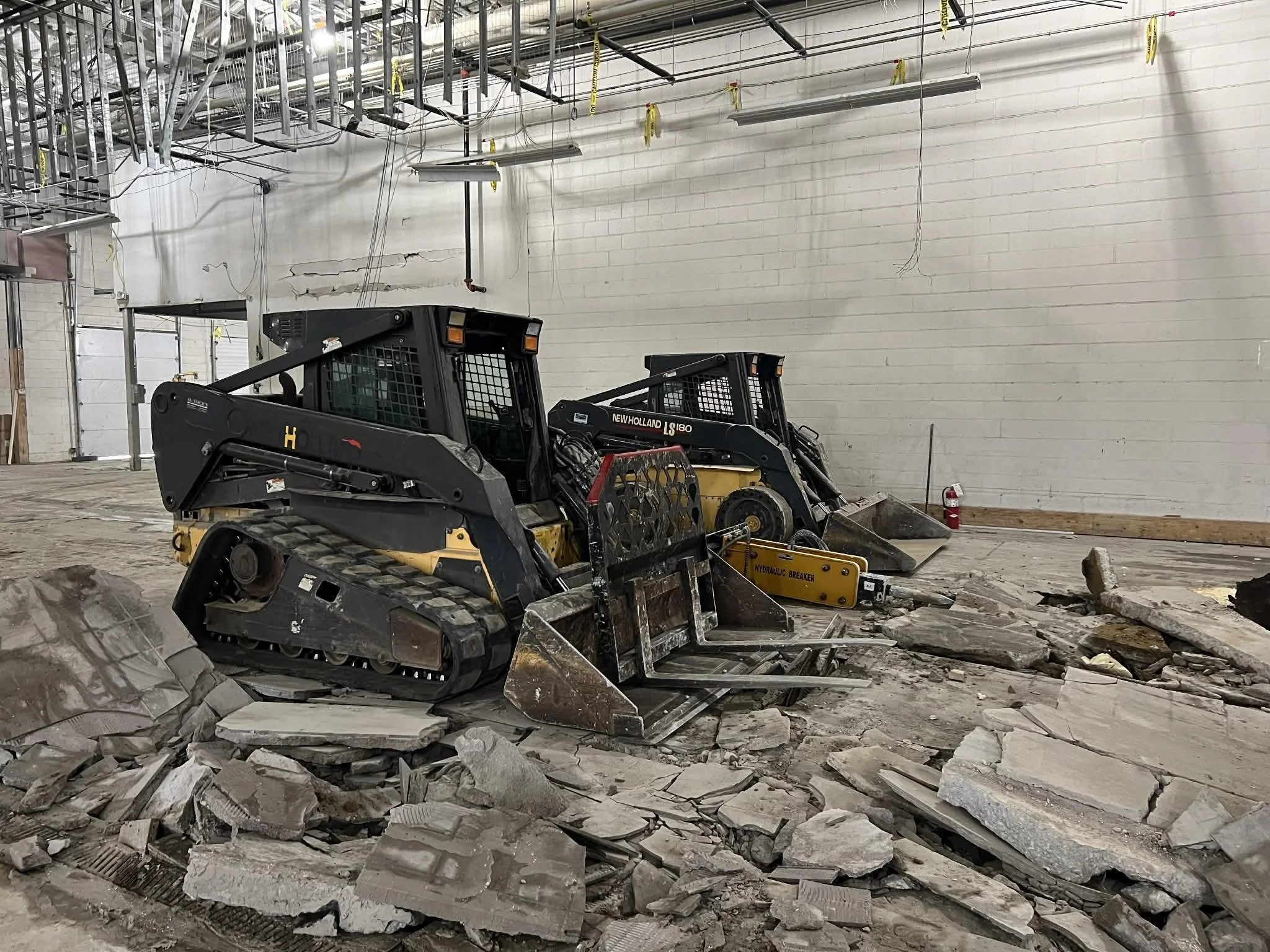 Construction site with two small bulldozers breaking concrete floor, with scattered debris, in an indoor utility or warehouse space