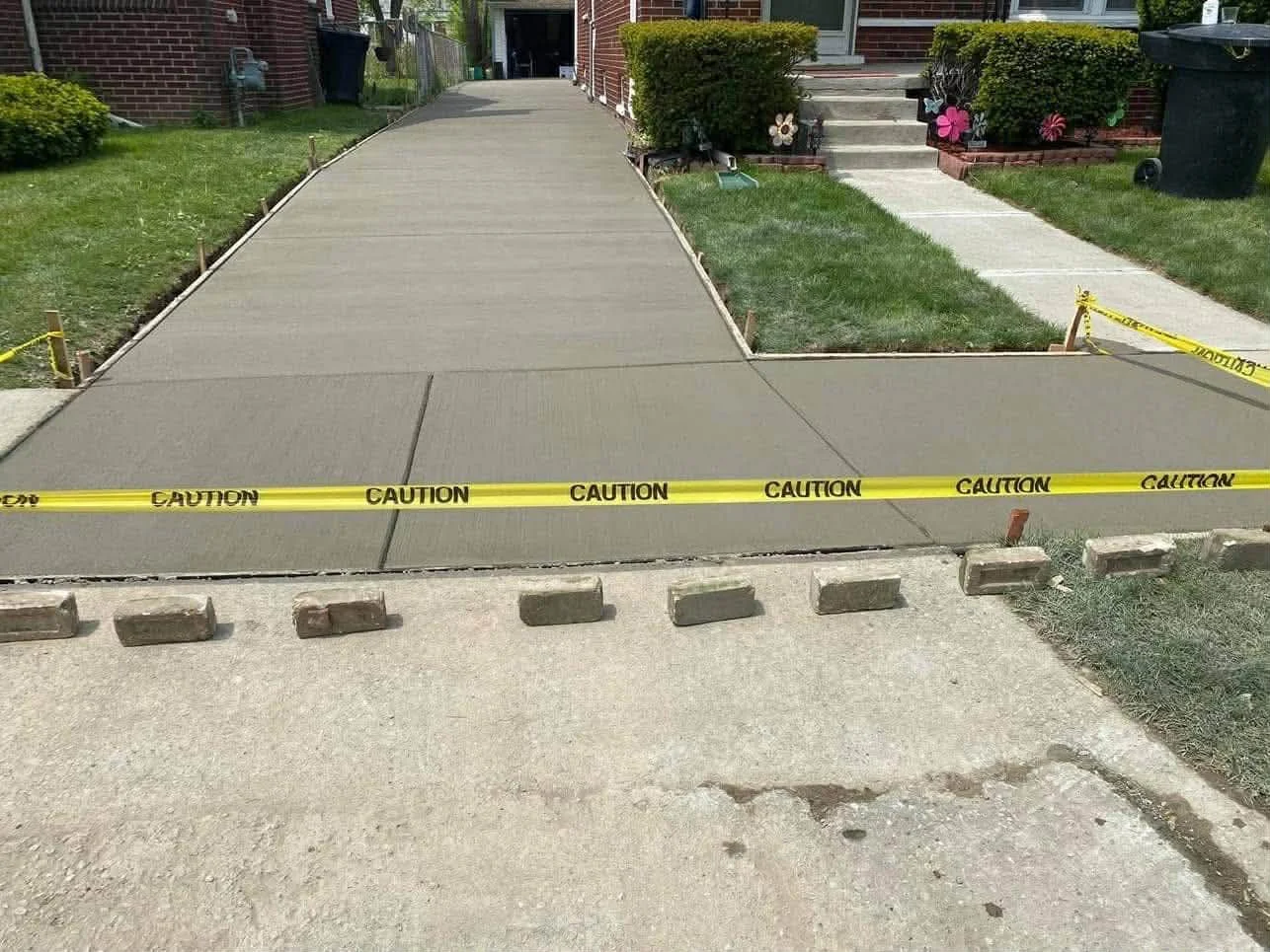 Concrete driveway and sidewalk in a residential front yard, with caution tape blocking the driveway and small concrete barriers at the entrance, and flower pots on the porch steps.