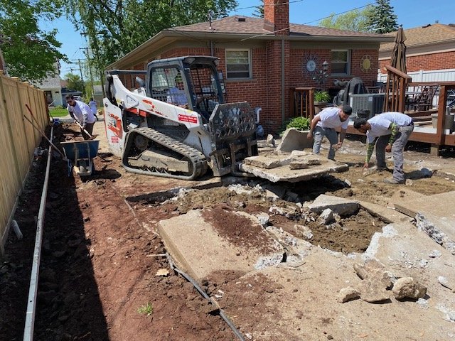 Three workers are involved in a backyard construction project, removing concrete slabs with a small skid-steer loader. They are working near a brick house with a deck, and there are tools and equipment around.