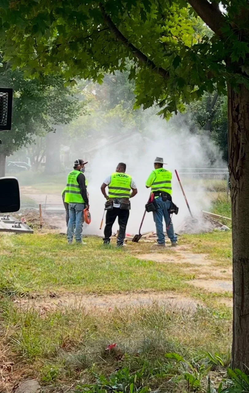 Construction workers in yellow vests working on a firebreak or clearing brush on a grassy area with smoke in the background and trees overhead.