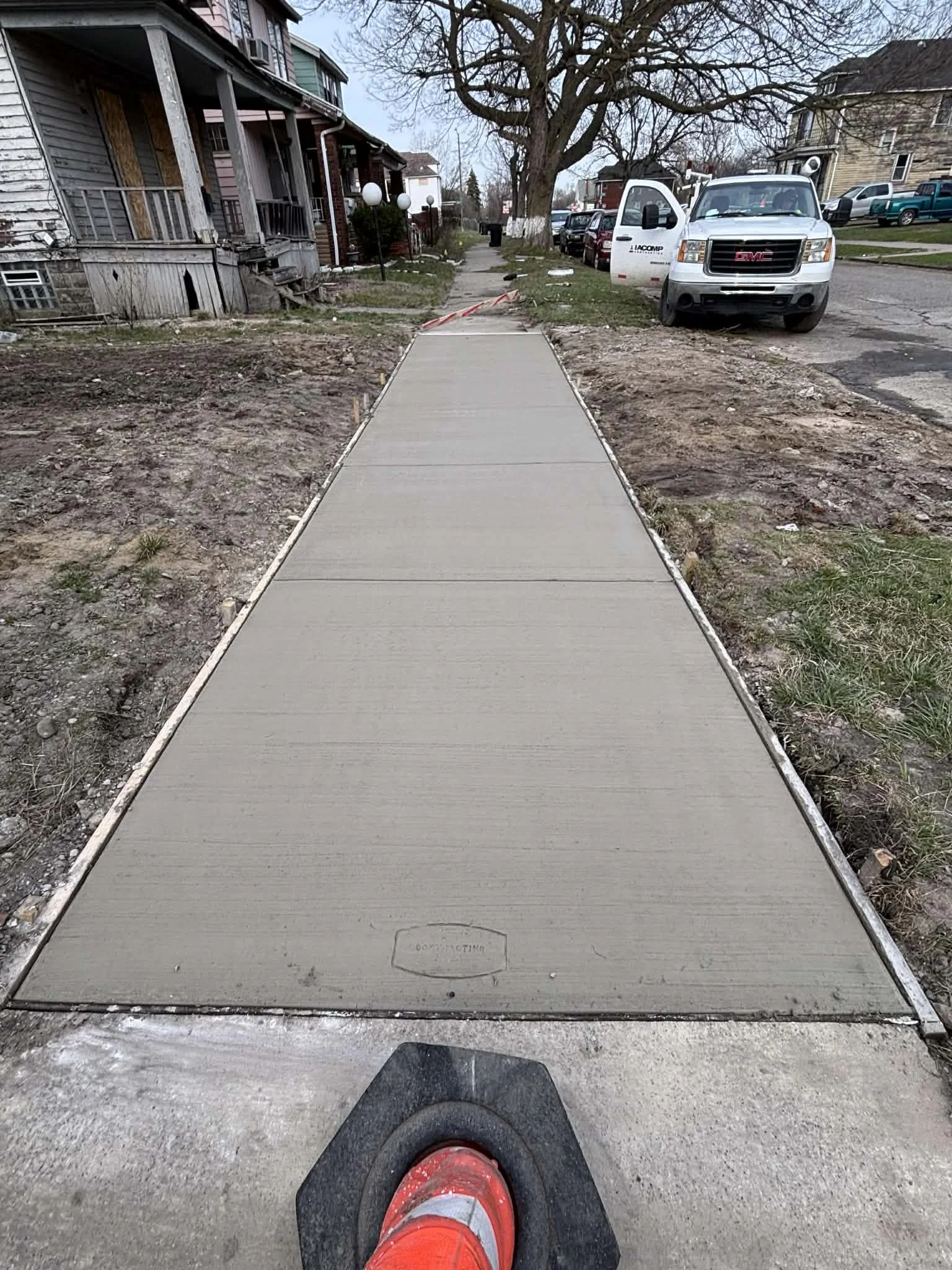 Freshly poured concrete sidewalk extending into a residential neighborhood with houses, parked cars, and leafless trees.
