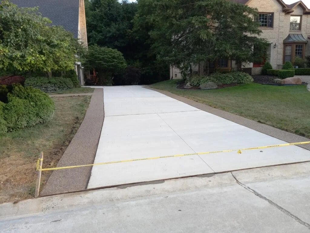 Concrete driveway with a yellow caution tape at the entrance, surrounded by grass, bushes, and residential houses in the background.