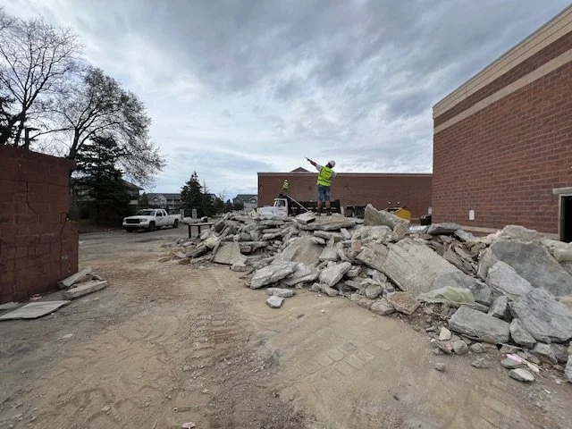 Two construction workers in safety vests and helmets working on debris and rubble near a brick building, with a cloudy sky overhead.