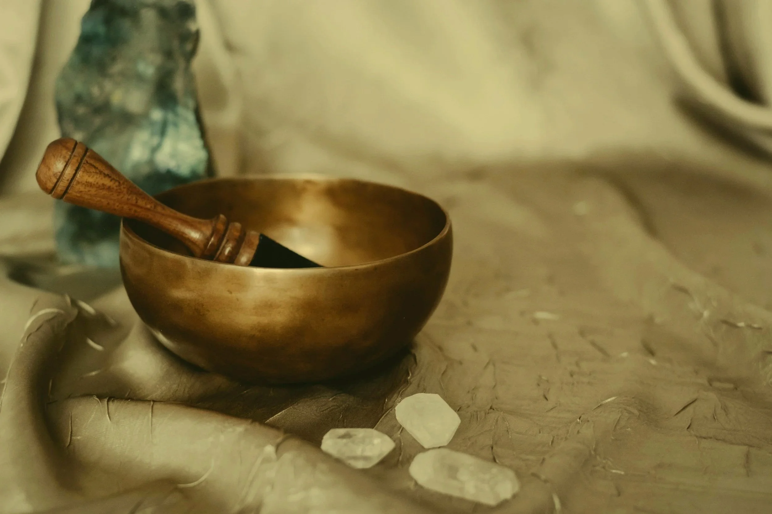 A wooden singing bowl with a mallet rests on a textured fabric surface, surrounded by three clear quartz crystals, with a blurred background.