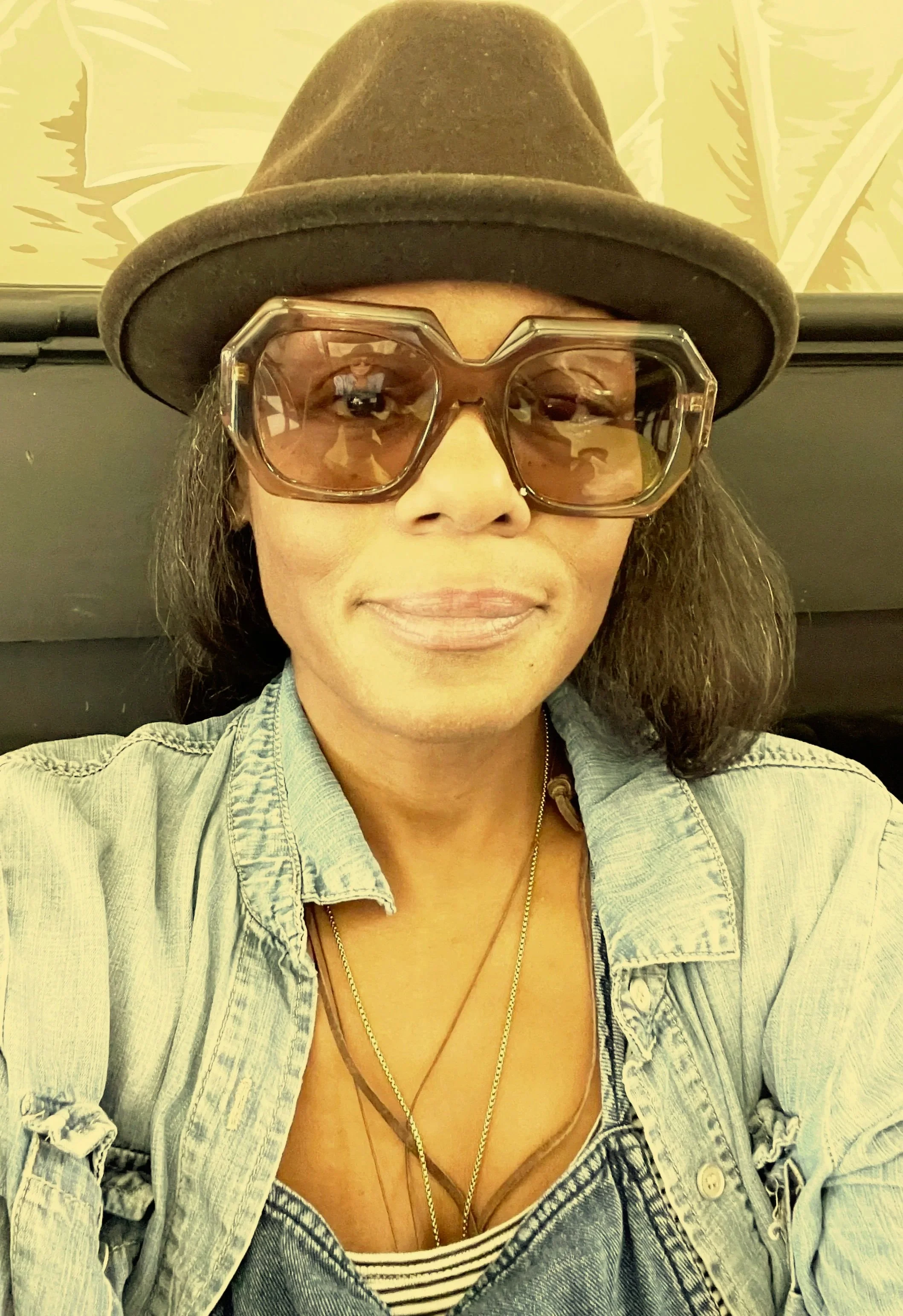 A woman wearing a brown hat, large tinted glasses, a light denim jacket, and layered necklaces, sitting in front of a leafy patterned wall.