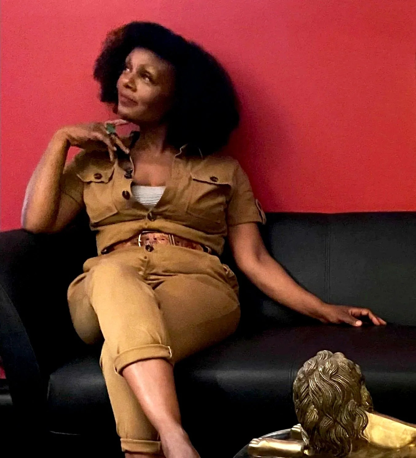 Woman with large curly hair sitting on a black couch against a red wall, wearing a tan outfit, relaxing with one arm on the couch and the other hand near her face.