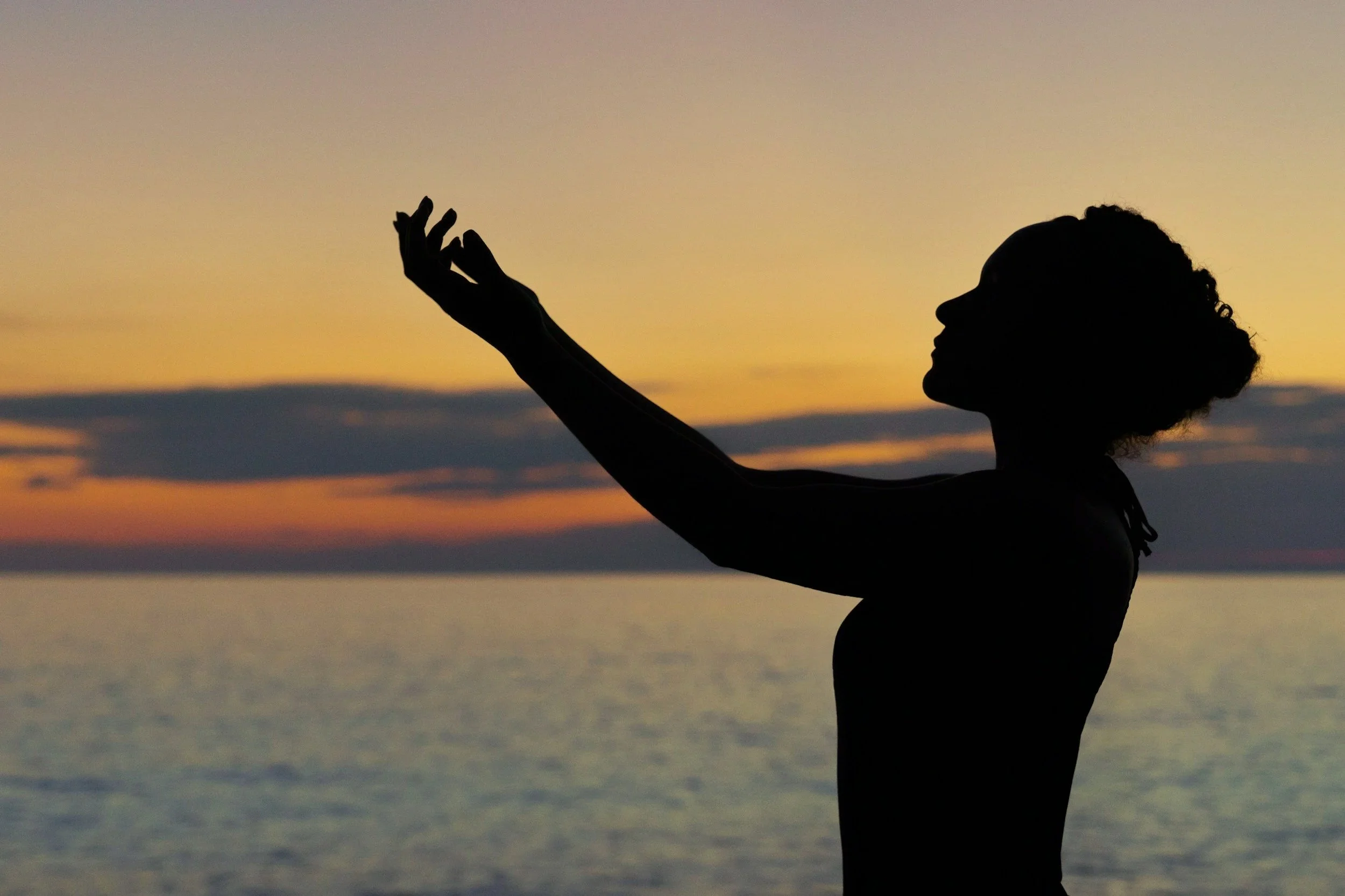 Silhouette of a woman stretching her arm at sunset by the water with a colorful sky in the background.