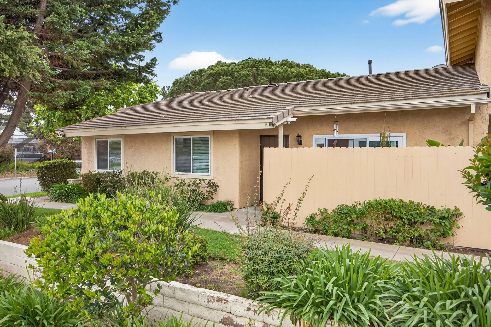 The photo shows the back view of a beige house with a sloped roof, a white wooden fence, and a garden with various green plants and bushes in front. There are tall trees and a blue sky with some clouds in the background.