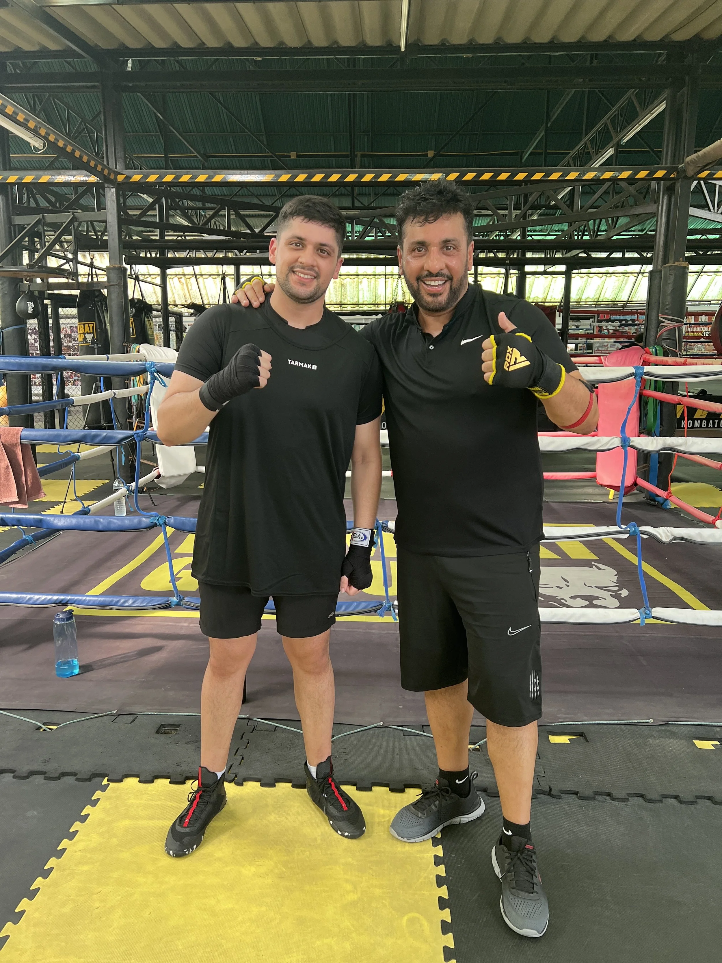 Two athletic men with dark hair and beards pose with fists raised in front of a boxing ring inside a gym. Both are wearing black athletic clothing and gloves, smiling at the camera.