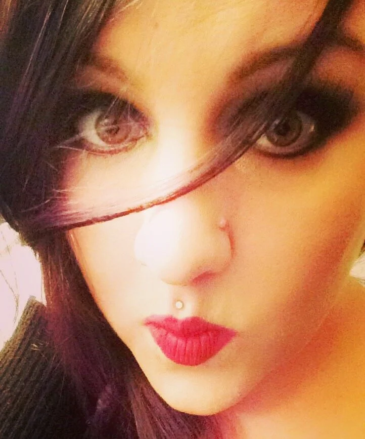 A close-up selfie of a woman with light skin, dark hair, bold red lipstick, and a nose piercing, with some hair partially covering her face, this is Jamie.