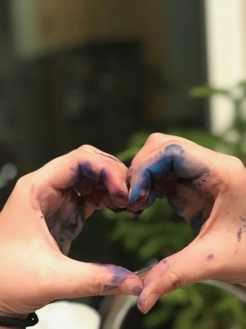Two hands forming a heart shape with fingers, covered in blue and purple ink or paint, against a blurry indoor background.