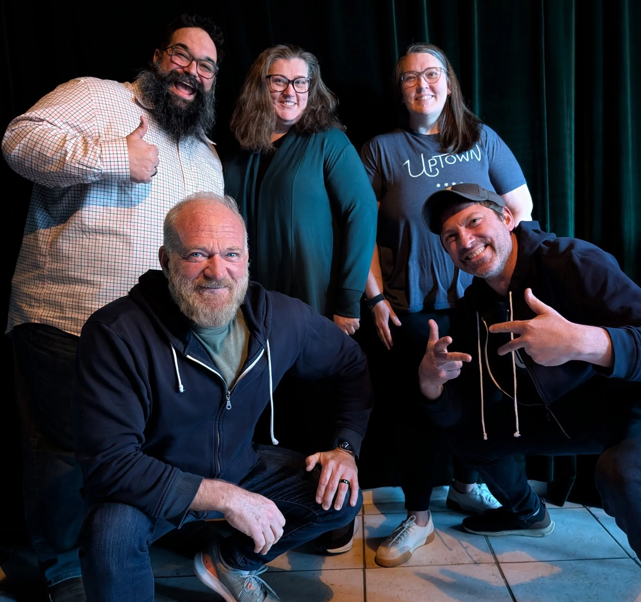 Group of five smiling people who run the Uptown Art Walk, two kneeling in front, three standing behind, posing for a photo indoors against dark curtains.