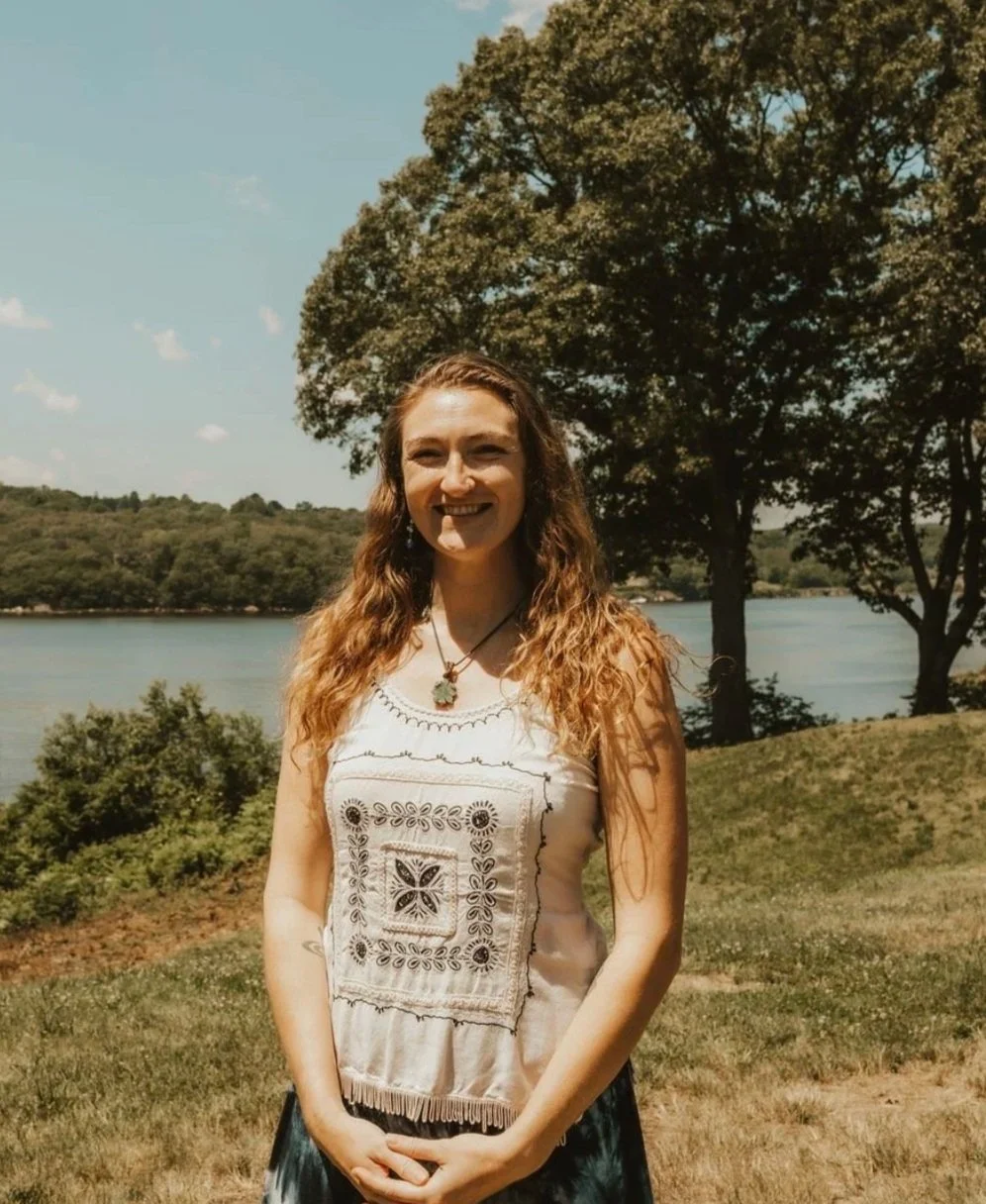 A woman standing outdoors near a lake, smiling at the camera. She has long, wavy hair and is wearing a white embroidered sleeveless top and a necklace. There are trees and grassy areas in the background under a partly cloudy sky.