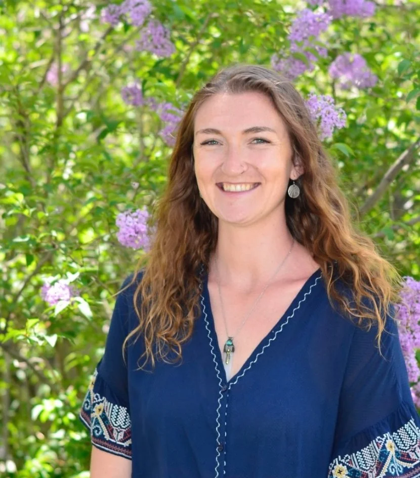 A woman with long, curly brown hair smiling outdoors in front of green foliage and purple flowers, wearing a blue top with embroidered sleeves.
