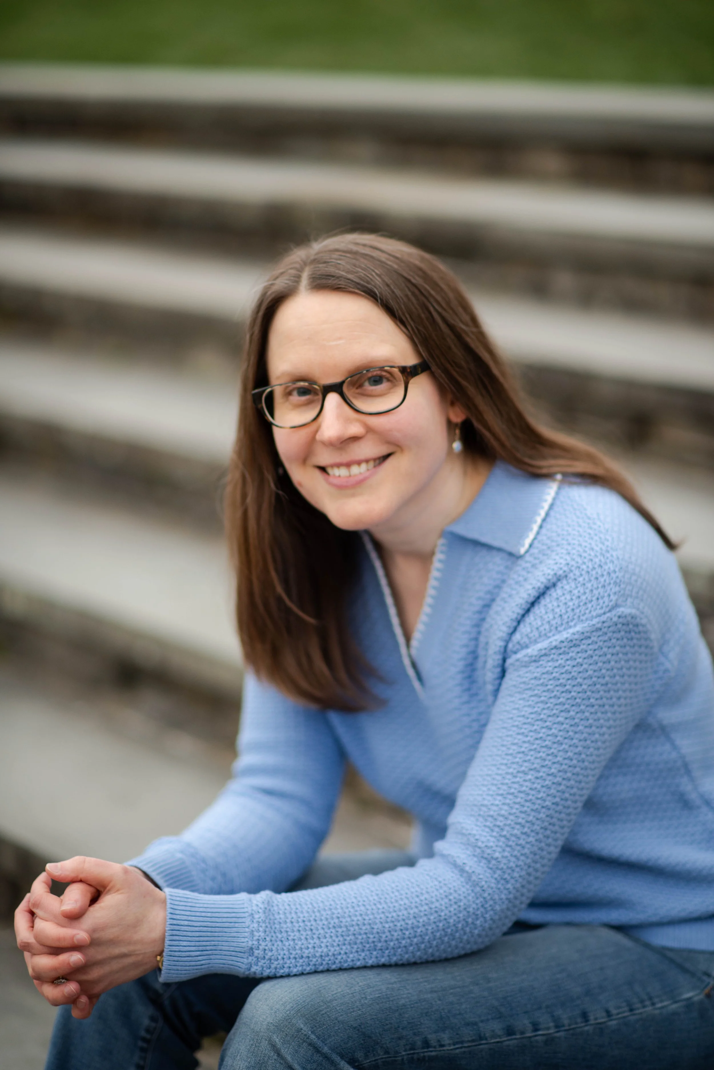 A woman with long brown hair, glasses, and a blue sweater sitting on outdoor steps, smiling.