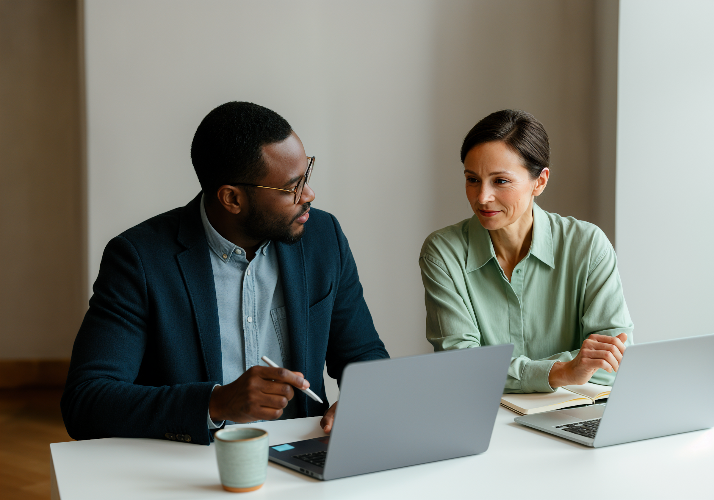 Two people, a man and a woman, sitting at a table with laptops, engaged in a discussion in a professional setting.