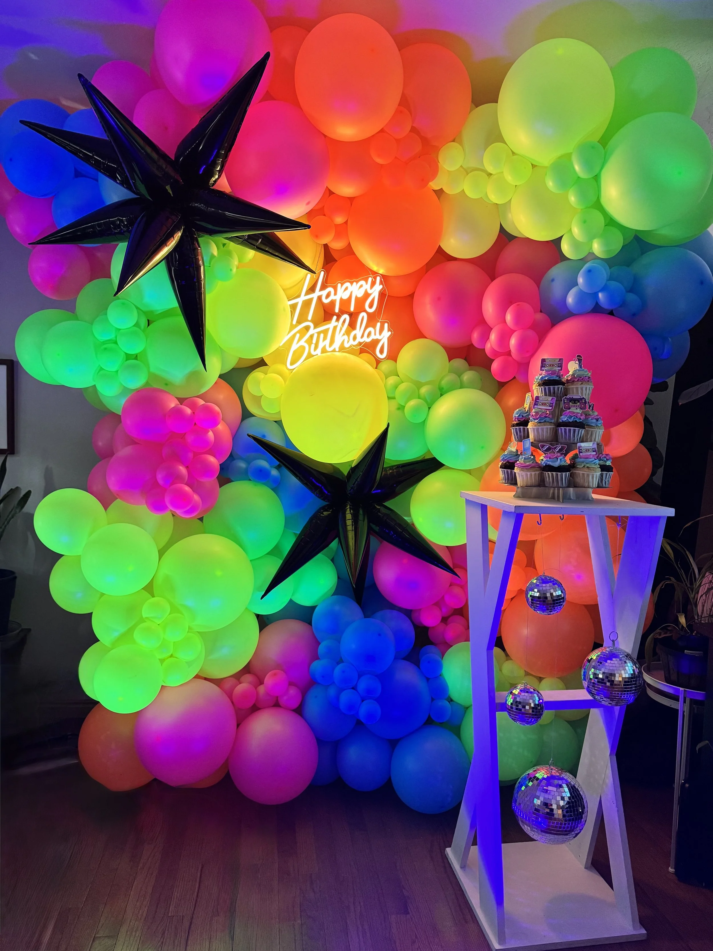 Colorful balloon wall with neon 'Happy Birthday' sign, black star-shaped balloons, and a white table holding cupcakes decorated for a birthday celebration, illuminated by disco balls.