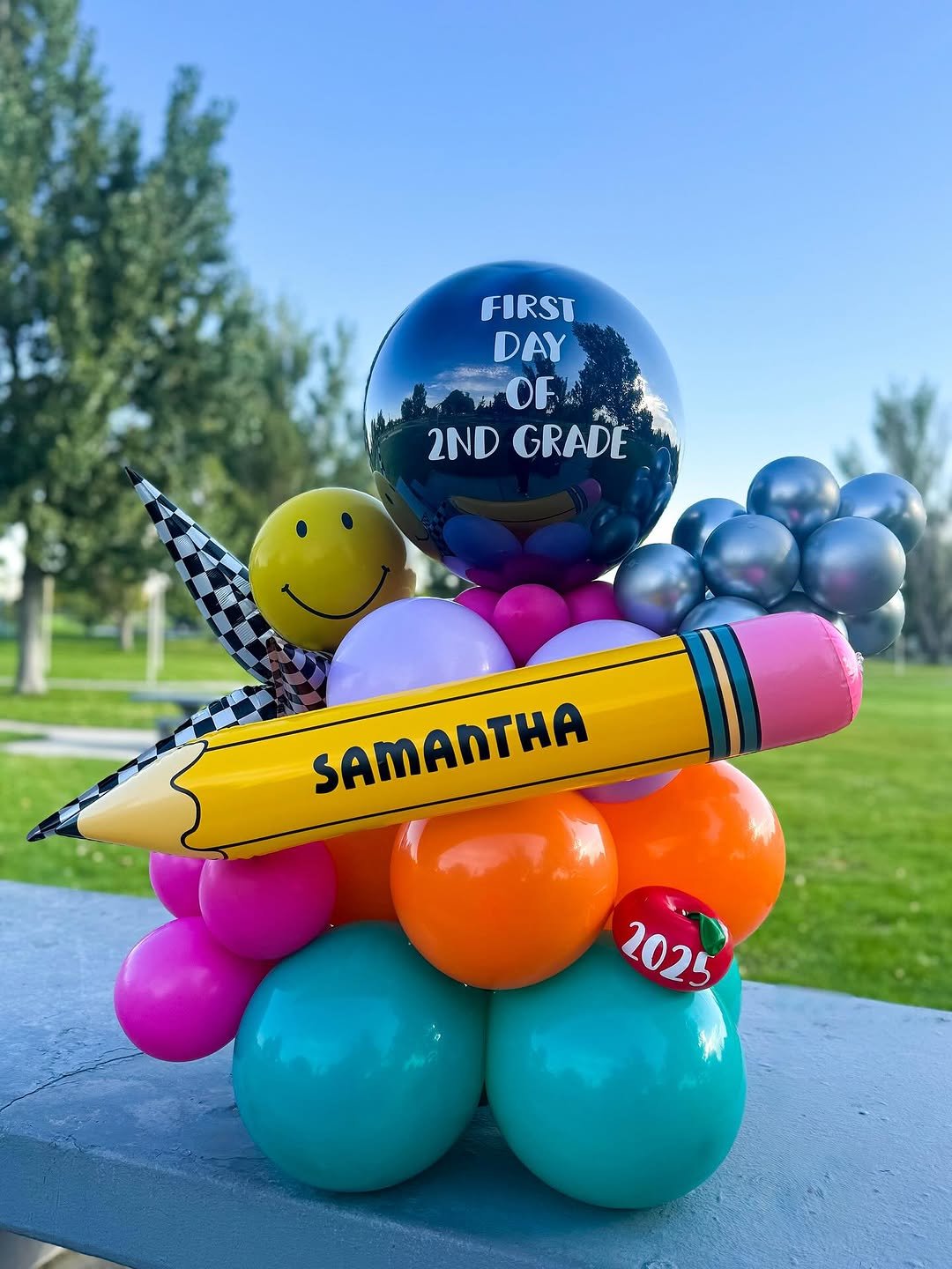 Balloon arrangement for a back-to-school celebration including a large black balloon with white text 'First Day of 2nd Grade', a yellow smiley face balloon, a large yellow pencil-shaped balloon with 'Samantha' written on it, a red apple balloon with 