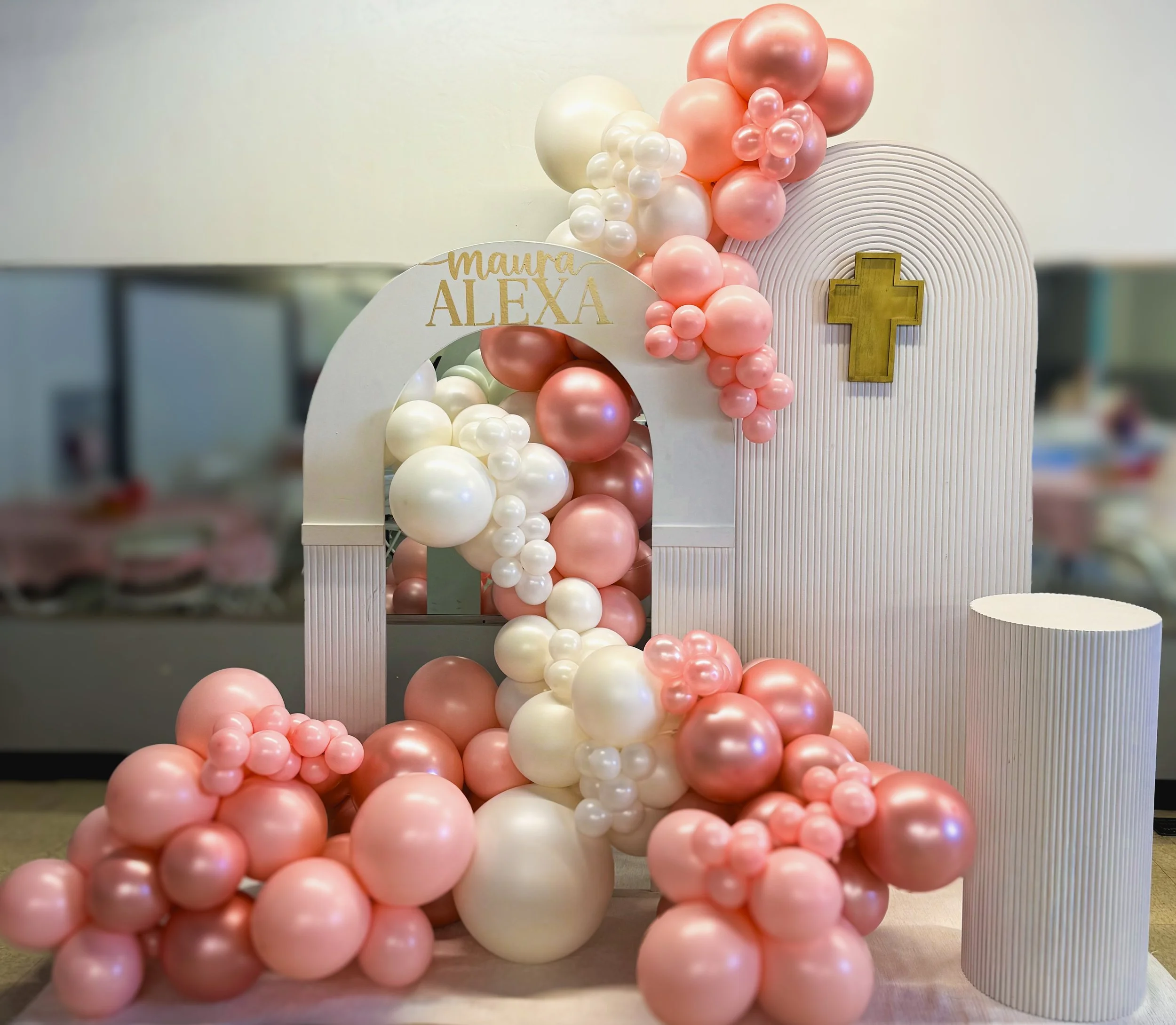 Decorative display of pink, white, and pearl-colored balloons arranged around a white arch with a gold cross and a sign that reads 'Maura Alexa,' set in an indoor space.