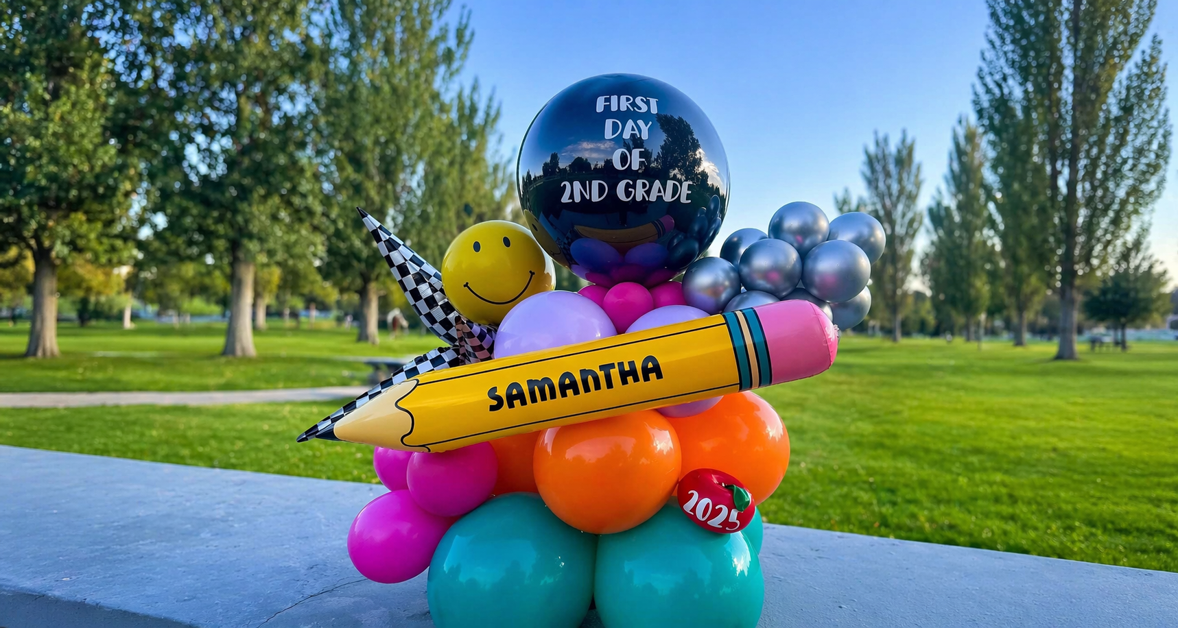 Colorful balloons and decorations, including a large black balloon with the words "First Day of 2nd Grade," a giant yellow pencil with the name "Samantha," a smiley face balloon, a checkered party hat, and a small red apple with "2025" on it, set up outdoors in a park with trees and green grass.