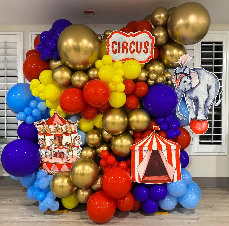 Colorful circus-themed balloon display with red, yellow, blue, purple, and gold balloons, along with circus-themed cutouts including an elephant holding a ball, a carousel with horses, and a red and white striped circus tent.