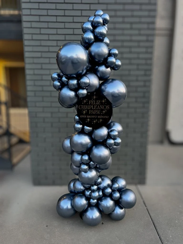 A balloon arrangement with metallic blue spheres of varying sizes, topped with a black circular sign in Spanish that says 'Feliz Cumpleaños Papá, eres nuestro universo'.