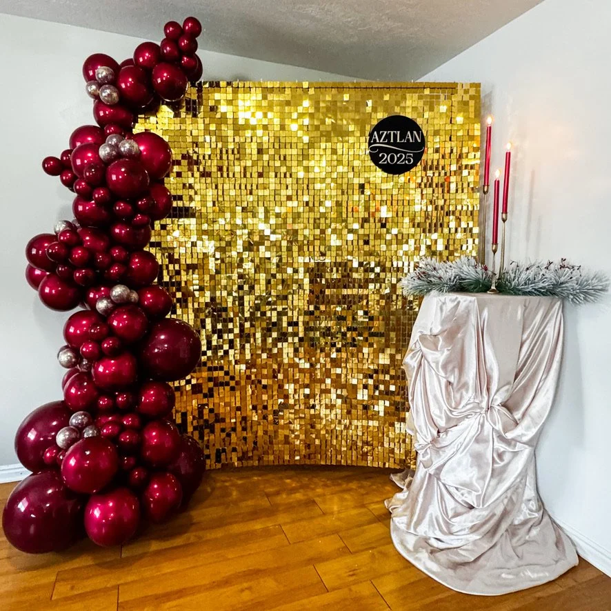 Gold sequin backdrop with red and silver balloons cluster on the left and a white fabric-covered table with candles and greenery on the right, celebrating AZTLAN 2025.