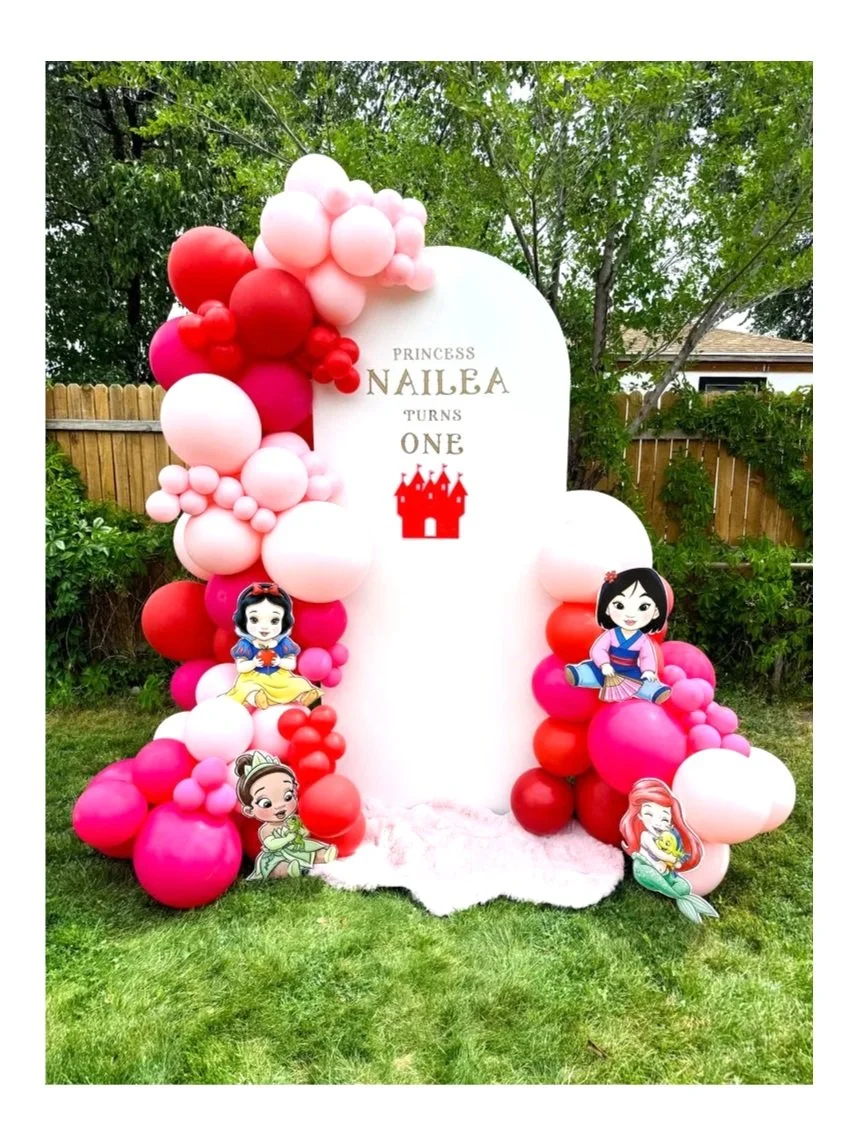 Birthday decoration with pink, white, and red balloons, Disney princess cutouts, and a white backdrop with a castle graphic, celebrating Nailea's first birthday in a backyard.